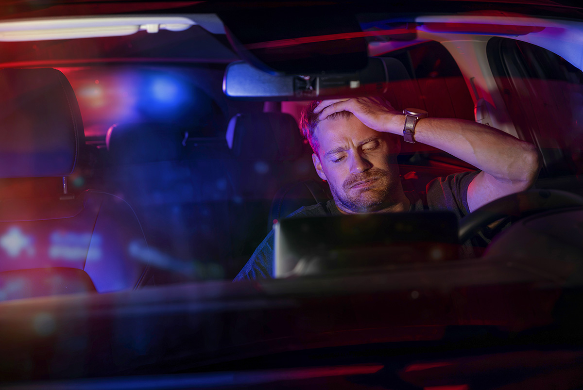 A man sitting in a car with red and blue police lights in the background as part of our series on signs of an impaired driver.