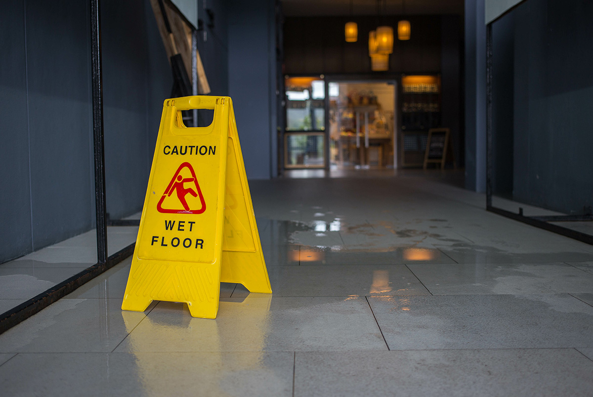 A yellow caution sign next to a puddle of water in a dimly lit, gray entryway leading to glass doors to demonstrate spill management.