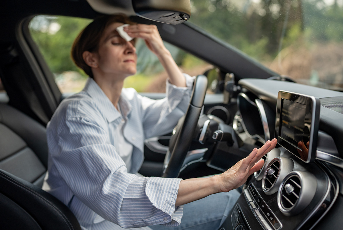 A woman in the driver seat holding a towel to her forehead and reaching a hand over the air conditioning vent to demonstrate heat exhaustion.