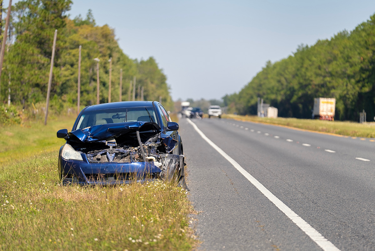 A broken down car on the side of the highway with cars approaching in the background.