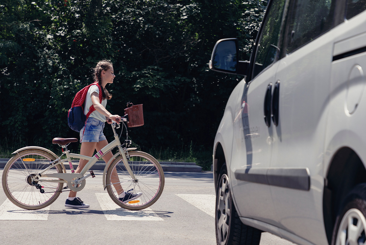 A little girl in a backpack walking her bike in front of a car at an intersection to illustrate back to school safety tips.