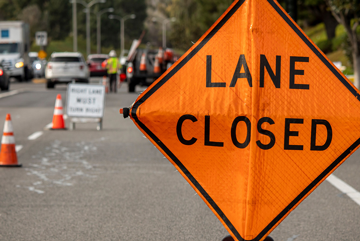 A close up of an orange Lane Closed construction zone sign with cars and workers blurry in the background.